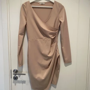 Beige omlottklänning från NLY One - Säljer en beige kortklänning från NLY One i omlottmodell med lång ärm och v-ringad hals. Klänningen har draperad detalj framtill som ger en snygg siluett. Tillverkad i ett mjukt och stretchigt material som sitter bekvämt på kroppen.