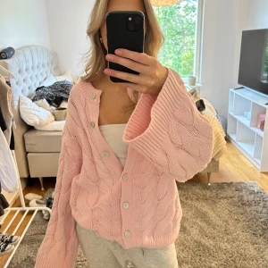 Rosa kabelstickad kofta med knappar - Supermysig rosa kofta med kabelstickat mönster och stora, vida ärmar. Koftan har v-ringning och knäpps med vita knappar framtill. Perfekt för lager-på-lager och chill dagar. Snygg att styla med både jeans och mjukisbyxor.