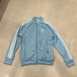 Blå Adidas ziptröja med vita ränder - Säljer en ljusblå Adidas ziptröja med klassiska vita ränder längs ärmarna och Adidas-logga på bröstet. Tröjan har lång ärm, ribbade muddar och dragkedja framtill. Perfekt för en sportig och chill stil. Använt 2 gånger. 