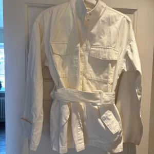 Vit jacka med bälte från NA-KD - Säljer en stilren vit jacka från NA-KD med bälte i midjan och stora bröstfickor. Jackan har lång ärm, knappar vid ärmslut och är gjord i bomull/jeans. 