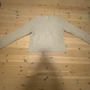 Beige stickad tröja från H&M - Mysig beige stickad tröja från H&M med kabelstickat mönster. Tröjan har rund halsringning, långa ärmar och ribbade muddar vid ärmslut och nederkant. Perfekt för lager-på-lager under kyliga dagar.