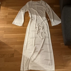 Vit långklänning i satin med knytband - Säljer en elegant vit långklänning i satin med långa utsvängda ärmar och smalt knytband i midjan. Klänningen har en enkel och stilren design med rund halsringning och diskret dragkedja bak. Den är lite genomskinlig och kan passa perfekt till semestern/stranden. Perfekt för dig som vill ha en clean och classy look.
