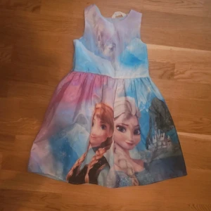 Frozen-klänning med motiv i pastell - Supersöt ärmlös klänning med motiv från Frozen i blå, lila och rosa toner. Klänningen har rund halsringning och utställd kjol, perfekt för dig som älskar Disney och vill sticka ut med färgglada vibes.