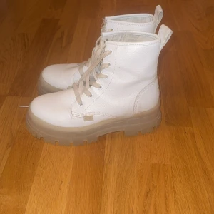 Buffalo vita boots med platåsula - Buffalo boots i vitt skinn med chunky, transparent platåsula. Klassisk snörning framtill och dragflik bak. Runda tår och robust design som ger en cool och edgy vibe. Perfekta för dig som vill sticka ut med dina skor.