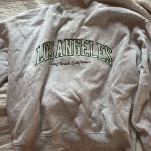 Grå sweatshirt Los Angeles tryck - Grå sweatshirt med grönt och svart Los Angeles-tryck på bröstet. Klassisk rund halsringning och ribbade muddar vid ärmslut och nederkant. Perfekt för en chill och avslappnad stil.
