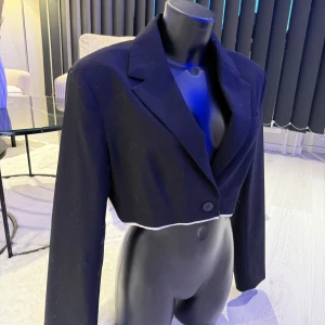 Svart croppad kavaj med knappar - Snygg svart croppad kavaj med klassisk krage och en knapp framtill. Långa ärmar med tre dekorativa knappar vid ärmslutet och vit kontrastkant nedtill. Perfekt för dig som vill ha en trendig och edgy look.