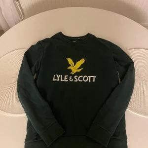 Mörkgrön sweatshirt från Lyle & Scott - Säljer en mörkgrön sweatshirt från Lyle & Scott med klassisk gul logga och vit text på bröstet. Tröjan har rund hals, långa ärmar och ribbade muddar. Perfekt för dig som gillar stilrena och sportiga plagg.