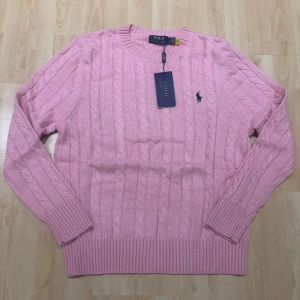 Rosa kabelstickad tröja Polo Ralph Lauren - Kabelstickad rosa tröja från Polo Ralph Lauren med klassisk rund halsringning och broderad marinblå logga på bröstet. Tröjan har långa ärmar och ribbade muddar för en snygg passform. Perfekt för dig som gillar preppy stil.