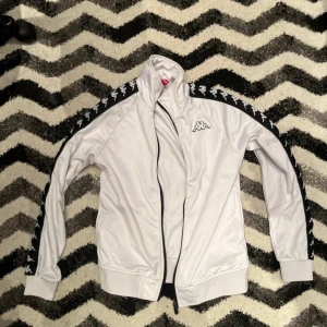 Vit track jacket från Kappa XS - Vit track jacket från Kappa med svarta detaljer och klassiska loggor längs ärmarna. Jackan har dragkedja framtill, hög krage och ribbade muddar. Tillverkad i 100% polyester för en sportig och clean look.