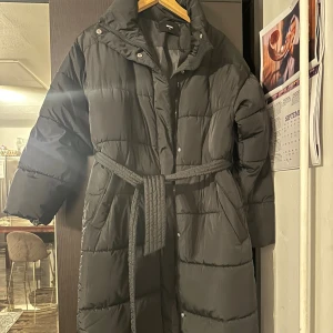 Svart lång pufferjacka med bälte - Lång svart pufferjacka från BikBok med quiltad design och bälte i midjan. Jackan har hög krage, dragkedja och tryckknappar framtill samt långa ärmar. Perfekt för kalla dagar när du vill hålla dig varm och snygg. Jackan har även slits där nere.