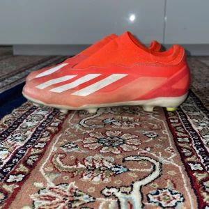 Adidas crazyfast laceless - Säljer ett par röda Adidas fotbollsskor med vita ränder på sidorna. Skorna har en strumpliknande passform utan snörning och är designade för att sitta tajt på foten. Yttersulan har fasta dobbar för spel på gräs eller turf. Perfekta för dig som vill ha stilrena och snabba skor på planen.