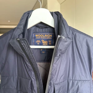 Mörkblå dunväst från Woolrich - Snygg mörkblå dunväst från Woolrich med hög krage och dragkedja framtill. Västen har två bröstfickor med lock och två sidofickor. Tillverkad i ett lätt och slitstarkt syntetmaterial. Nypris kostar denna 3.400kr säljer för 1.200kr!! 