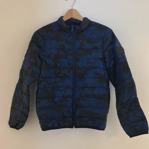 Blå camo pufferjacka med dragkedja - Säljer en blå pufferjacka med camouflagemönster och hel dragkedja framtill. Jackan har ståkrage, elastiska muddar vid ärmslut och en patch på ena ärmen. Perfekt för kyliga dagar och riktigt snygg streetstil.