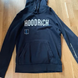 Mörk blå hoodie från Hoodrich med tryck - Mörk blå hoodie från Hoodrich med stor vit logga framtill och grafiskt tryck i vitt och blått på ryggen. Tröjan har huva med snörning, känguruficka och långa ärmar. Perfekt streetwear-vibe och riktigt skön passform.