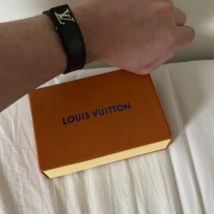 Louis Vuitton svart armband  - Snyggt svart armband från Louis Vuitton med ikonisk LV-logo i silver och klassiskt monogrammönster. Armbandet är stilrent och har exklusiv känsla, perfekt för dig som gillar lyxiga accessoarer. Levereras i originalask med dustbag och certifikat.