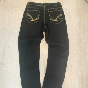 Svarta DNY jeans med broderade bakfickor - Säljer ett par svarta jeans från DNY med snygga kontrastsömmar och unika broderade detaljer på bakfickorna. Klassisk femficksmodell med raka ben och normal passform. Jeansen är tillverkade i ett slitstarkt jeanstyg som passar perfekt till din streetwear-look.