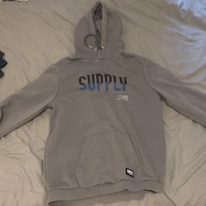 Supply and demand hoodie - Grå hoodie från Supply and Demand med svart och blått tryck på bröstet. Klassisk känguruficka framtill, justerbar huva med snörning och ribbade muddar. Perfekt för chill dagar och streetstyle. Mjuk och skön känsla.