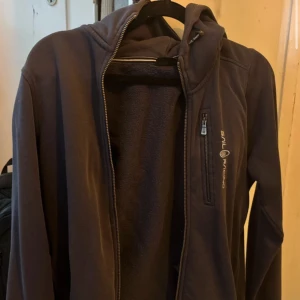 Mörkblå fleecekofta Sail Racing - Mörkblå fleecekofta från Sail Racing med huva och hel dragkedja. Jackan har en bröstficka med dragkedja och logga på bröstet. Mjuk insida i fleece och sportig look, perfekt för kyliga dagar.