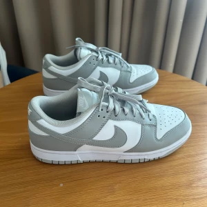 Nike Dunk Low grå/vita sneakers - Nike Dunk Low sneakers i stilren grå och vit färg. Klassisk siluett med lågt skaft, snörning och perforerad tå. Ovandel i syntet och skinn, med vit bas och ljusgrå overlays samt matchande grå swoosh. Perfekta för dig som gillar clean och modern streetstyle.