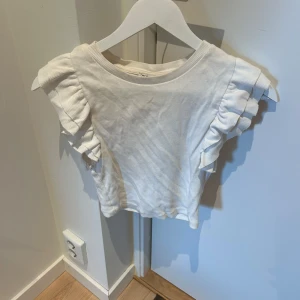 Vit volangtopp från Zara - Supersöt vit topp från Zara med ribbad struktur och volangdetaljer på axlarna. Toppen är ärmlös och har en rund halsringning. Perfekt för dig som gillar en feminin och trendig stil.