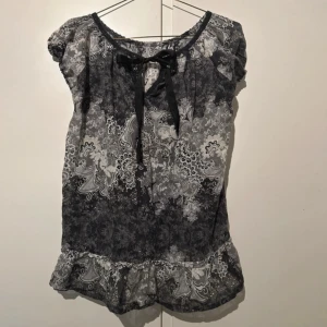 Mönstrad blus med rosett från Odd Molly - Supersöt blus från Odd Molly i svart och grått med paisleyinspirerat mönster. Blusen har korta ärmar, volangkant nertill och en snygg rosett vid halsringningen. Tunn och luftig i bomull, perfekt för dig som gillar detaljer och unik stil.