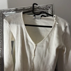 Vit ribbad kofta från Zara - Superfin vit ribbad kofta från Zara med v-ringning och knappar framtill. Långa ärmar med snygga volangdetaljer vid ärmsluten. Perfekt att slänga över ett linne eller t-shirt för en chill look. I S men passar XS