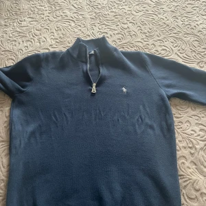 Mörkblå stickad tröja Polo Ralph Lauren - Mörkblå stickad tröja från Polo Ralph Lauren med halv dragkedja och broderad logga på bröstet. Tröjan har hög krage och är långärmad, perfekt för kyligare dagar. Klassisk och stilren design som funkar till det mesta.