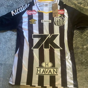 Santos FC randig fotbollströja Umbro - Santos FC matchtröja från Umbro med svartvita vertikala ränder, klubbmärke på bröstet och flera sponsorloggor. Tröjan har korta ärmar, gul detalj vid kragen och är tillverkad i lätt, ventilerande polyester. Perfekt för dig som älskar brasiliansk fotboll.