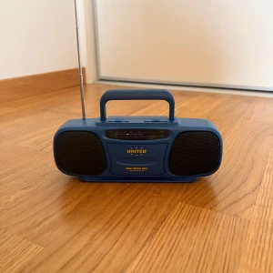 Mini Boom Box FM Radio Receiver - Snygg och portabel blå FM-radio från United, modell Mini Boom Box. Utrustad med två högtalare, teleskopantenn och praktiskt bärhandtag. Volym- och tuningreglage på ovansidan. Fint skick utan synligt slitage, perfekt för picknick eller resor.