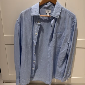 Ljusblå skjorta från H&M i linne - Ljusblå skjorta från H&M i relaxed fit med klassisk krage, bröstficka och knäppning framtill. Tillverkad i svalt linne, perfekt för en chill och stilren look. Långärmad modell med knappar vid ärmslut.