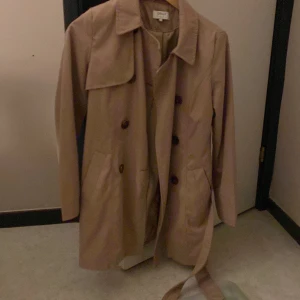 Beige trenchcoat från ONLY XS - Snygg beige trenchcoat från ONLY i storlek XS. Jackan har klassisk dubbelknäppning, bälte i midjan och stora knappar. Perfekt för dig som vill ha en stilren och tidlös look. Långärmad och med krage för extra stilpoäng.