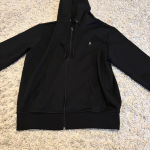 Svart hoodie från Polo Ralph Lauren - Svart hoodie från Polo Ralph Lauren med dragkedja, huva med snörning och klassisk logga broderad på bröstet. Tröjan har två fickor framtill och är tillverkad i mjuk bomull. Perfekt för en chill och stilren look.