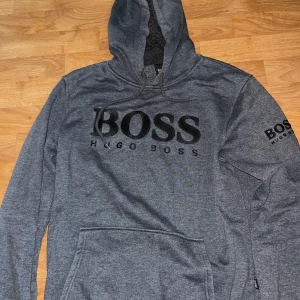 Grå hoodie från Hugo Boss med logga - Snygg grå hoodie från Hugo Boss med stor svart BOSS-logga på bröstet och mindre logga på ärmen. Tröjan har huva med snörning och en stor magficka. Perfekt för dig som gillar stilrena och bekväma plagg med premiumkänsla.