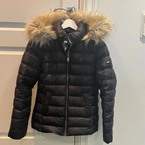 Svart pufferjacka Tommy Jeans - Svart pufferjacka från Tommy Jeans med huva och snygg fuskpäls runt kragen. Jackan har quiltad design, dragkedja framtill och logga på ärmen. Perfekt för kalla dagar och ger en cool streetstyle-vibe.