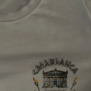 Vit Casablanca t-shirt med tryck - Vit t-shirt från Casablanca med stort grafiskt tryck på ryggen och mindre tryck på bröstet. Trycket har antika motiv, statyer, eld och texten 'Équipement Sportif'. Klassisk passform och rund hals. Tillverkad i 100% bomull.