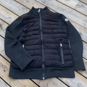 Moncler Cardigan - Svart Cardigan från Moncler med quiltad framsida och två dragkedjeförsedda fickor. Västen har en hög krage och Moncler-logga på axeln. Materialet är en mix av dun och polyester, vilket gör den både lätt och varm. Perfekt för lager-på-lager under kyliga dagar.