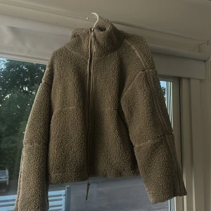 Beige teddyjacka med dragkedja - Mysig beige teddyjacka med hög krage och hel dragkedja framtill. Jackan har en boxig passform och markerade sömmar längs ärmar och kropp. Perfekt för kyliga dagar när du vill vara både varm och snygg.
