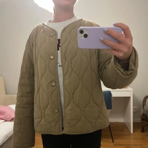 Beige quiltad jacka med knappar - Quiltad beige jacka med vågigt mönster, rund halsringning och knäppning framtill med fyra knappar. Jackan har en loose passform och är långärmad, perfekt för lager-på-lager under höst och vår. Tillverkad i mjukt bomullstyg för en chill och avslappnad vibe.