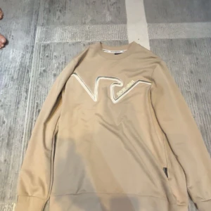 Beige sweatshirt från Emporio Armani - Snygg beige sweatshirt från Emporio Armani med broderad logga framtill i vitt. Tröjan har rund halsringning och långa ärmar, perfekt för en stilren och avslappnad look. Tillverkad i mjuk bomullsmix som känns skön mot huden.aldrig använd