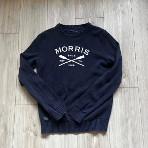 stickad tröja från Morris  - Snygg mörkblå stickad tröja från Morris med vit logga och text framtill. Tröjan har rund halsringning, långa ärmar och ribbade muddar vid ärmslut och nederkant. Perfekt för dig som gillar klassisk och stilren design.