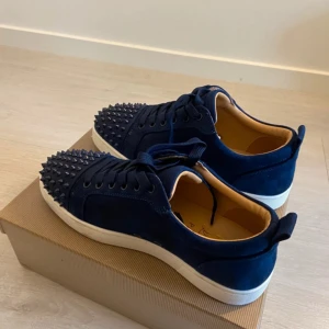 Blå mocka sneakers från Louboutin - Säljer ett par mörkblå sneakers från Christian Louboutin i mocka med coola nitar på tån och klassisk röd sula. Skorna har snörning och vit platt sula, samt en lyxig känsla med beige innersida. Perfekt för dig som vill sticka ut med din stil.