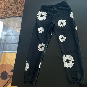 Svarta mjukisbyxor Denim Tears M - Svarta mjukisbyxor från Denim Tears med vita blommiga tryck över hela byxorna. Byxorna har resår i midjan med snörning och muddar vid bensluten. Perfekta för en chill och avslappnad stil.