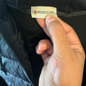 Svart cardigan från Moncler - Säljer en svart pufferjacka från Moncler med quiltad framsida, huva med dragsko och mjuka ärmar. Jackan har dragkedja framtill och två fickor. Klassisk Moncler-logga på ärmen och stilren design som funkar året runt.