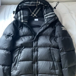 Svart pufferjacka från Burberry - Svart pufferjacka från Burberry med klassiskt rutigt foder i huvan. Jackan har dragkedja och knappar framtill, två snedställda fickor med dragkedja och en patch med Burberry London England på ärmen. Snygg och varm modell med justerbar huva.