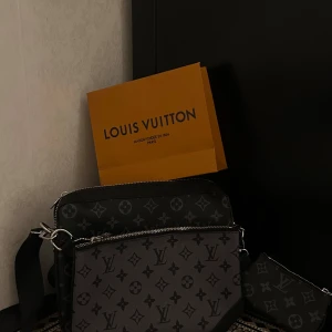 Svart axelväska monogram - Säljer en snygg svart axelväska med klassiskt monogrammönster i grått och svart. Väskan har flera fack, silverfärgade detaljer och justerbar axelrem. Tillverkad i exklusivt skinn och perfekt för dig som vill ha en stilren och lyxig accessoar.