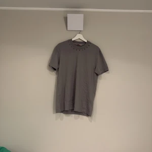 Grå t-shirt med nitar från Valentino - Snygg grå/gön t-shirt från Valentino med coola guldfärgade nitar runt halsringningen. Klassisk passform och tillverkad i mjuk bomull. Perfekt för dig som vill ha en stilren men edgy look.