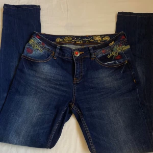Desigual broderade jeans med detaljer - Mörkblå jeans från Desigual med färgglada broderier av fjärilar och blommor, samt en röd tofs på bakfickan. Strl 31.