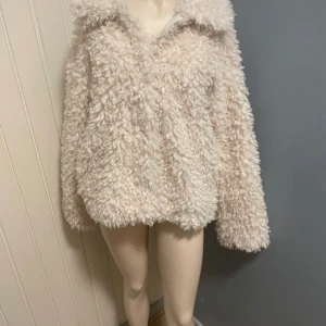 Beige teddyjacka med krage - Supermjuk och fluffig teddyjacka i beige med stora lockar och bred krage. Jackan har en loose passform och långa ärmar, perfekt för dig som vill ha en cozy och trendig look. Materialet är pälsimitation som känns riktigt gosigt.