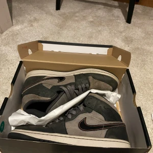 Nike Air Jordan 1 Mid grå/svart - Säljer ett par Nike Air Jordan 1 Mid i grått och svart med orange detaljer. Skorna har klassisk snörning, svart swoosh med orange söm och är tillverkade i mocka och skinn. Perfekta för dig som gillar streetwear och vill ha en ikonisk sneaker i garderoben.