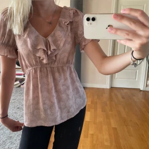 Ljusrosa blus med volang - Supersöt ljusrosa blus med mönster och volangdetaljer vid v-ringningen. Korta ärmar med lätt volang och resår i midjan för en feminin siluett. Perfekt för dig som gillar söt och trendig stil. Storlek S!